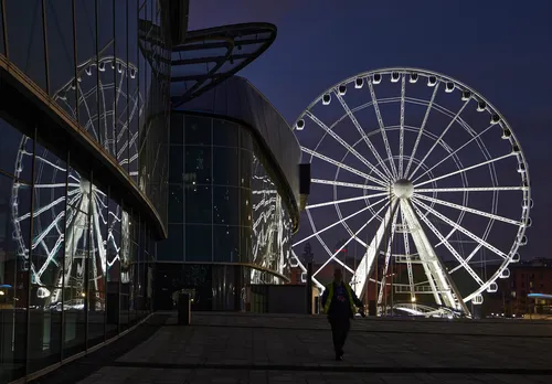 The image is an urban photograph taken in Liverpool, United Kingdom, during the evening in autumn. The main subject is the Liverpool Wheel, which is brightly illuminated and prominently reflected in the glass façade of a modern building. The reflections emphasize the circular structure of the wheel, creating a striking visual effect against the darkening sky. The scene captures the architectural details of both the wheel and the adjacent contemporary building, contributing to Liverpool's cityscape. The photograph highlights the interplay of light, glass, and structural design in an urban autumn setting, with the Liverpool Wheel providing a recognizable reference point within the city.